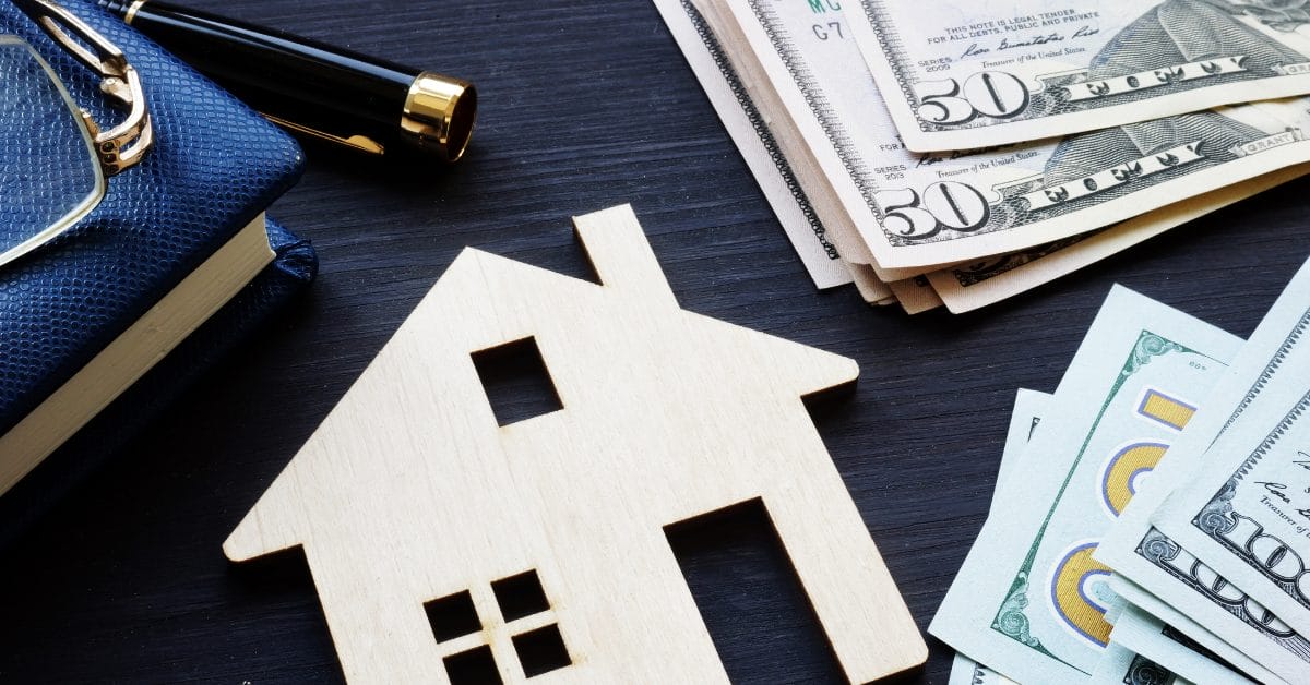 Wooden house on table with hundred dollar bills money and journal pen and glasses